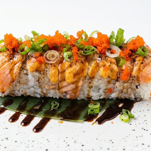 Sushi roll with seared salmon, orange tobiko, green onions, and sauce on a banana leaf.