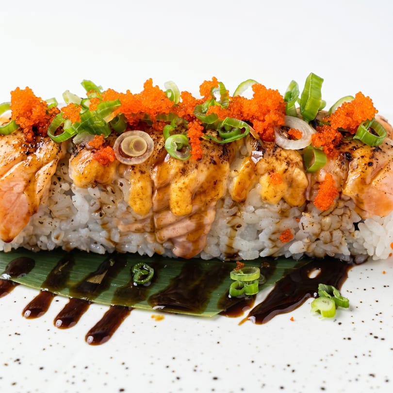 Sushi roll with seared salmon, orange tobiko, green onions, and sauce on a banana leaf.