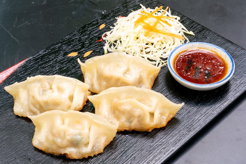 Four golden-brown dumplings, shredded cabbage, and a small bowl of dipping sauce on a black plate.