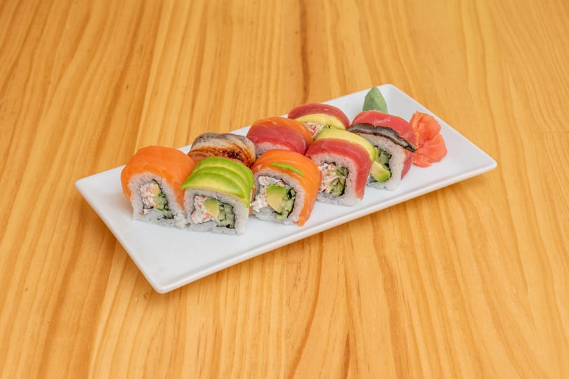 A white rectangular plate holds ten colorful sushi rolls, with ginger and wasabi on a wooden table.
